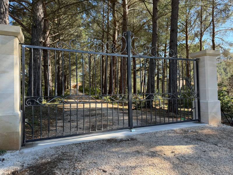 Portail battant en fer forgé traditionnel avec volutes ornementales, installé entre deux piliers en pierre de taille dans une propriété boisée à Aix-en-Provence.